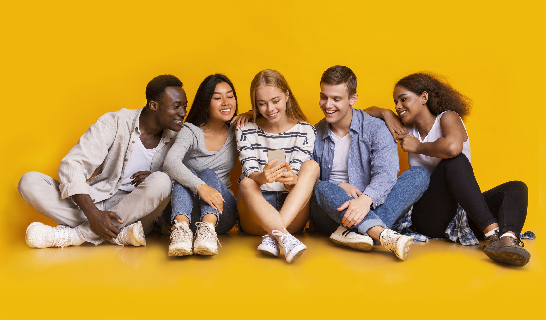 Teens lifestyle. Multiracial group of teenagers sitting on floor and looking at smartphone screen, panorama